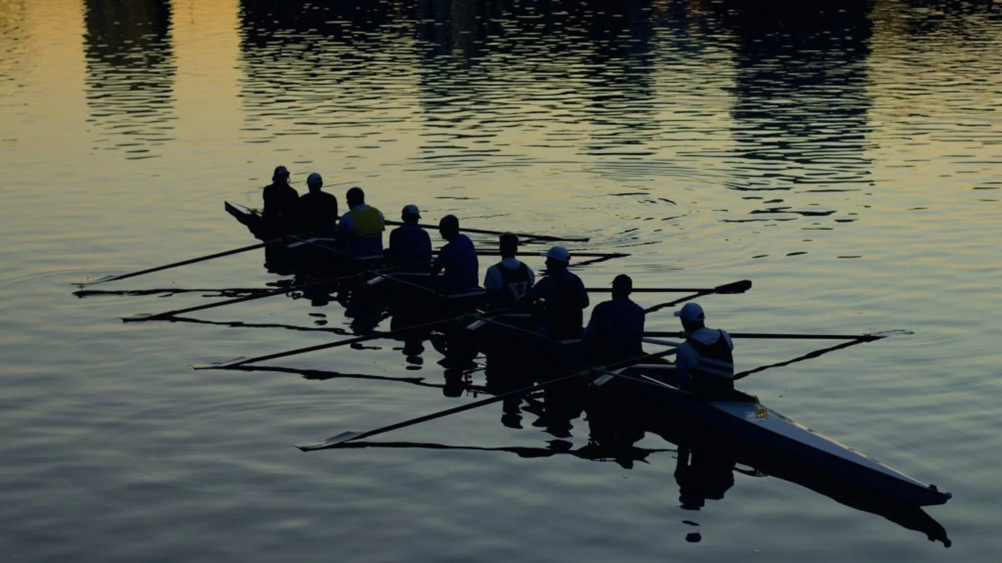 A rowing eight in silhouette on gold-lit water, every oar entering the water on the same stroke.